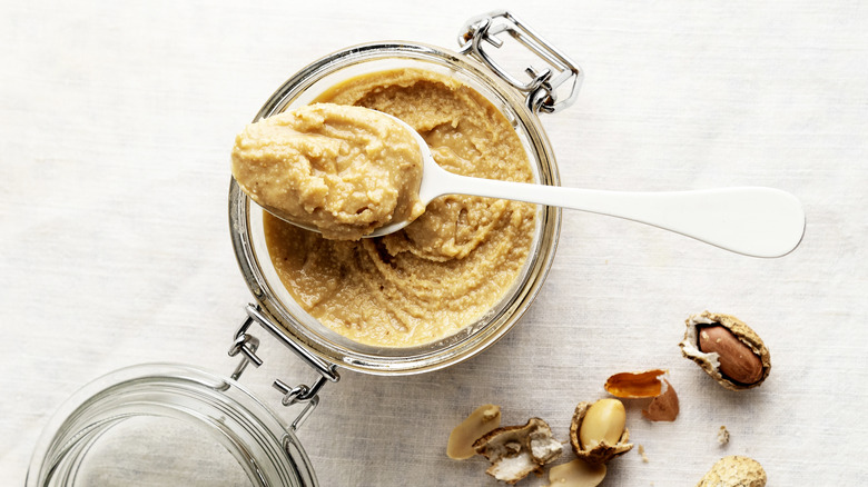 Peanut butter in glass jar next to  cracked peanuts