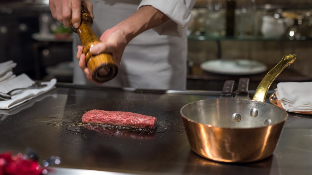 chef's hand grinding salt onto kobe beef on griddle in kitchen