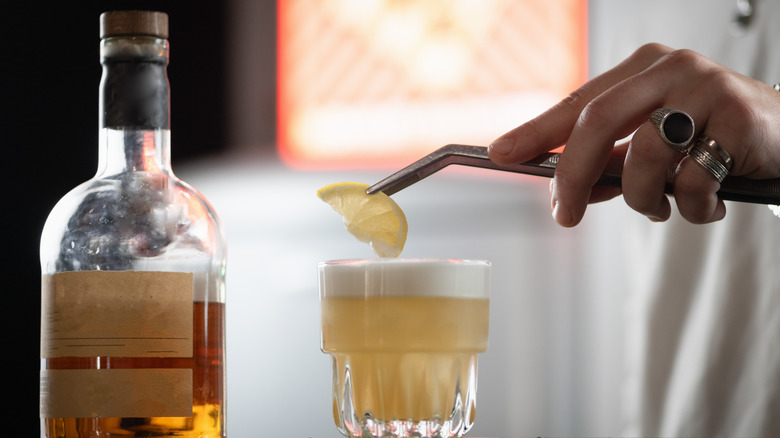 Hand using tongs to place lemon slice in pineapple whiskey sour.