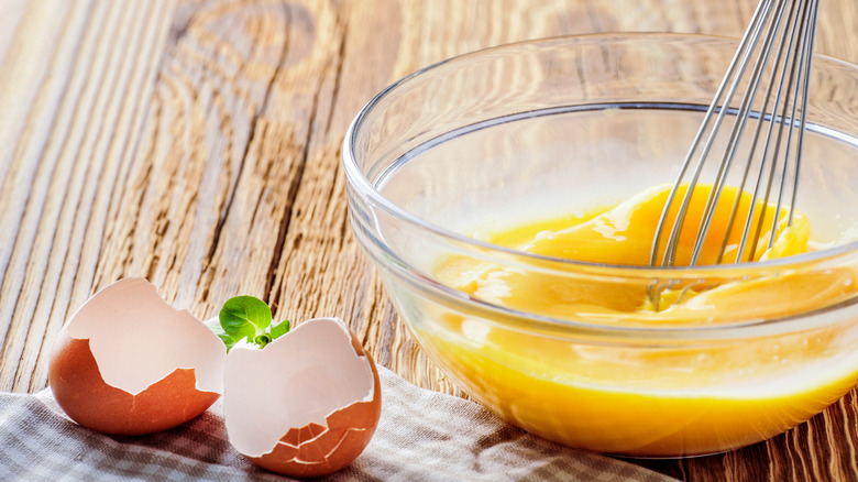 Whisk beating eggs in a clear bowl with egg shells on a towel